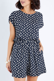Model Andrea wearing size 10 of The Printed Tie Waist Playsuit in colour Multiprint (Black), from the front, cropped image