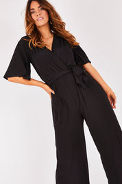 Model Laura wearing size 12 of The Bell Sleeve Jumpsuit in colour Black (Black), from the front, cropped image