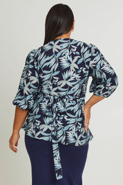 Model Ashleigh wearing size 16 of The Printed Blouson Sleeve Wrap Blouse in colour Navyleaf (Blue), from the back