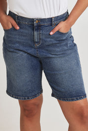Model Terri-Leigh wearing size 14 of the Daily Dose Longline Denim Shorts in colour Denim (Blue), from the front, cropped image