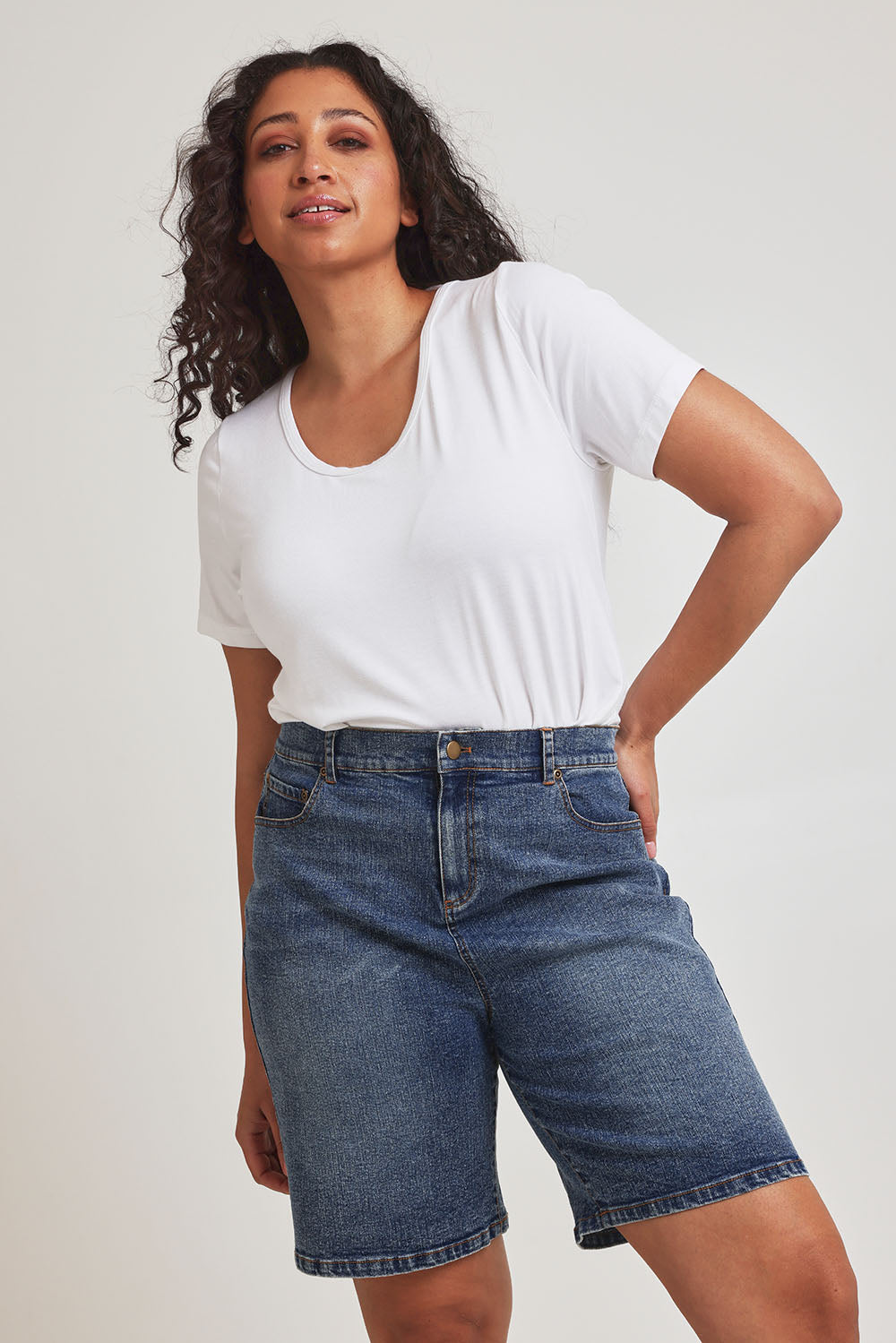 Model Terri-Leigh wearing size 14 of the Daily Dose Longline Denim Shorts in colour Denim (Blue), from the front, cropped image