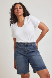 Model Terri-Leigh wearing size 14 of the Daily Dose Longline Denim Shorts in colour Denim (Blue), from the front, cropped image