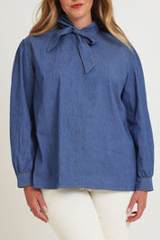 Model Chanel wearing size 16 of the Take A Bow Chambray Blouse in colour Demin (Blue), from the front, cropped image