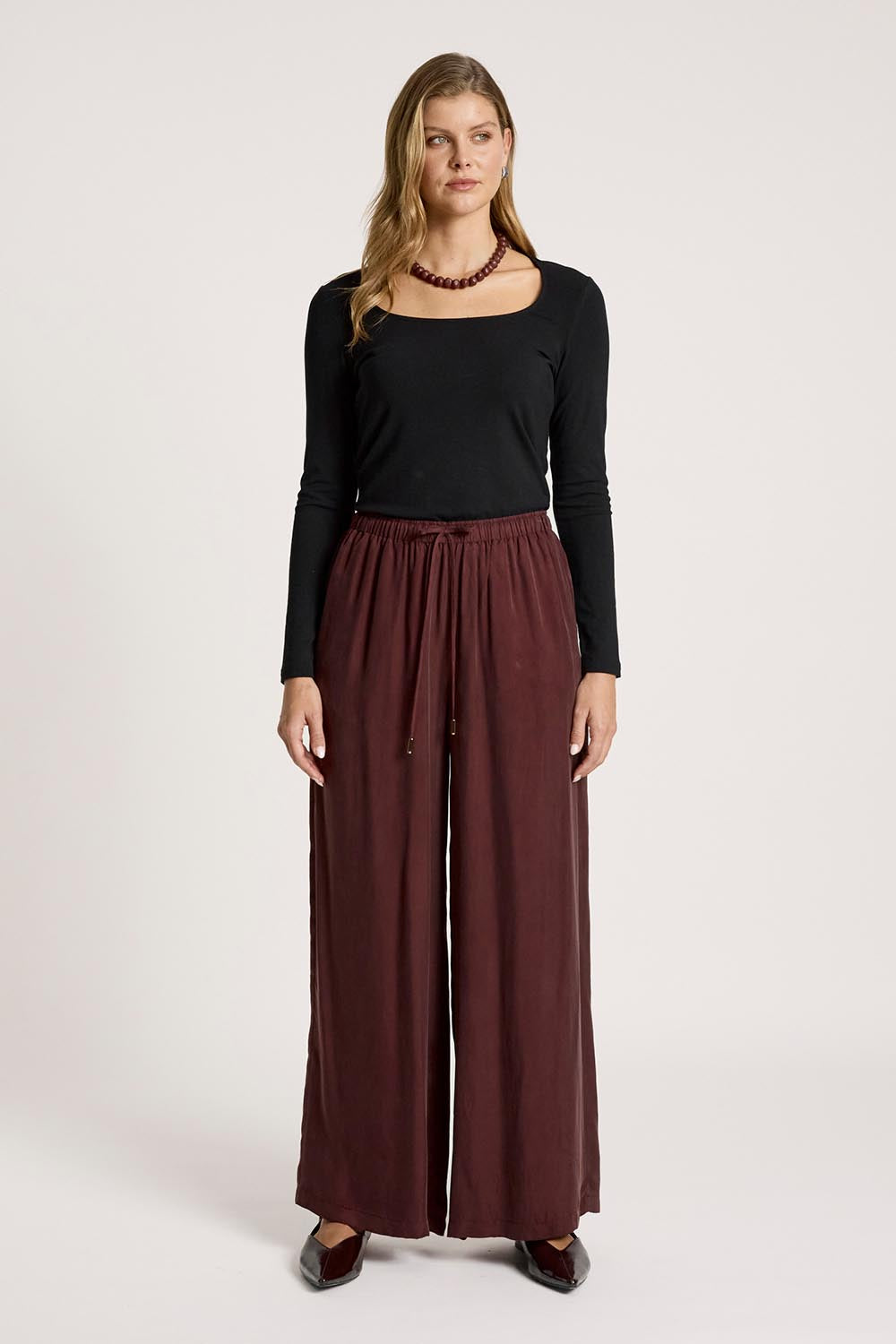 A model wearing the Calista Pant in colour Mahogany (Red), from the front, showing the whole outfit