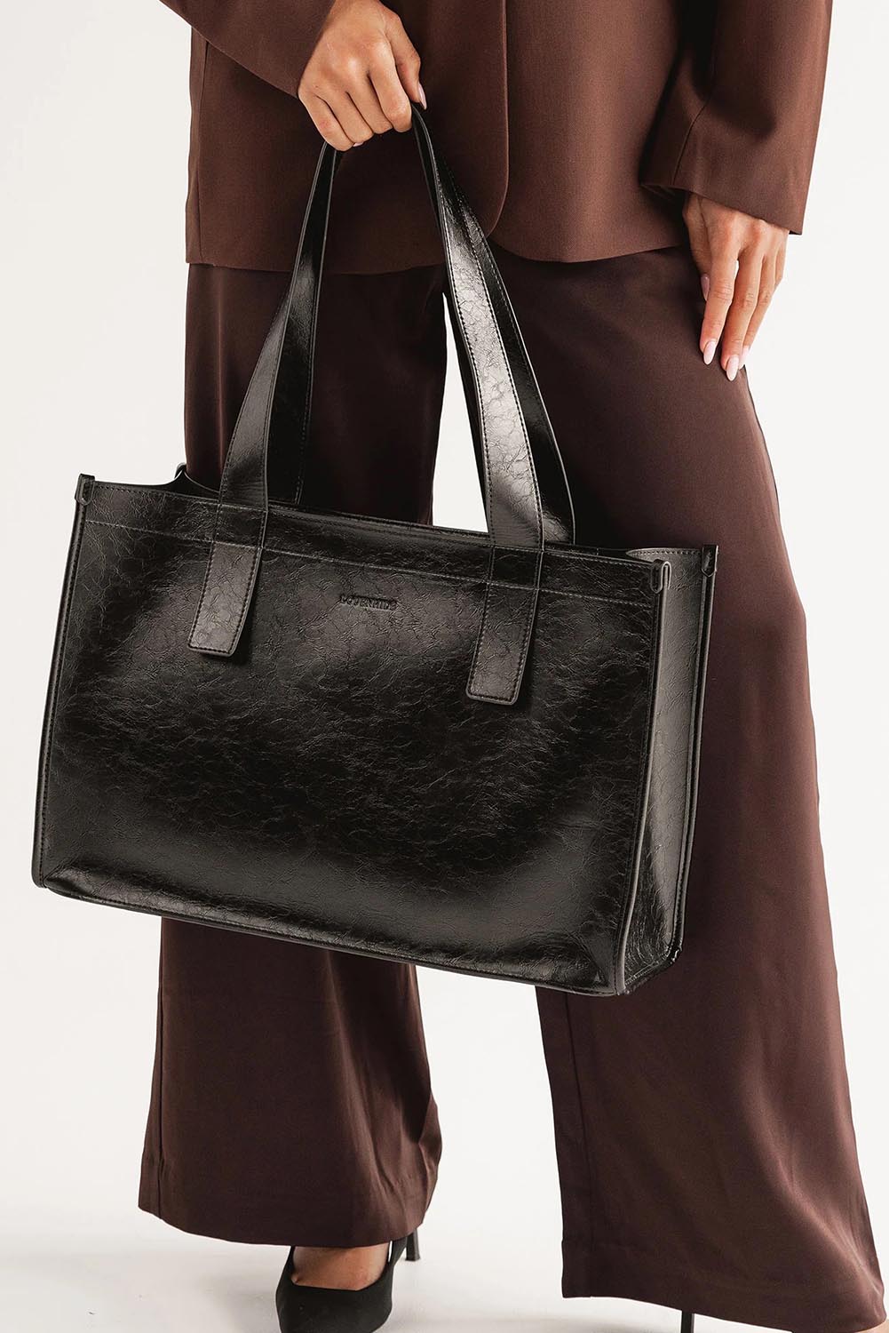 A model wearing the Riley Tote Bag in colour Blackpaten (Black), from the front, cropped image