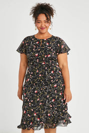 Model Alexis wearing size 16 of The Printed Chiffon Lined Dress in colour Botanical (Black), from the front, cropped image