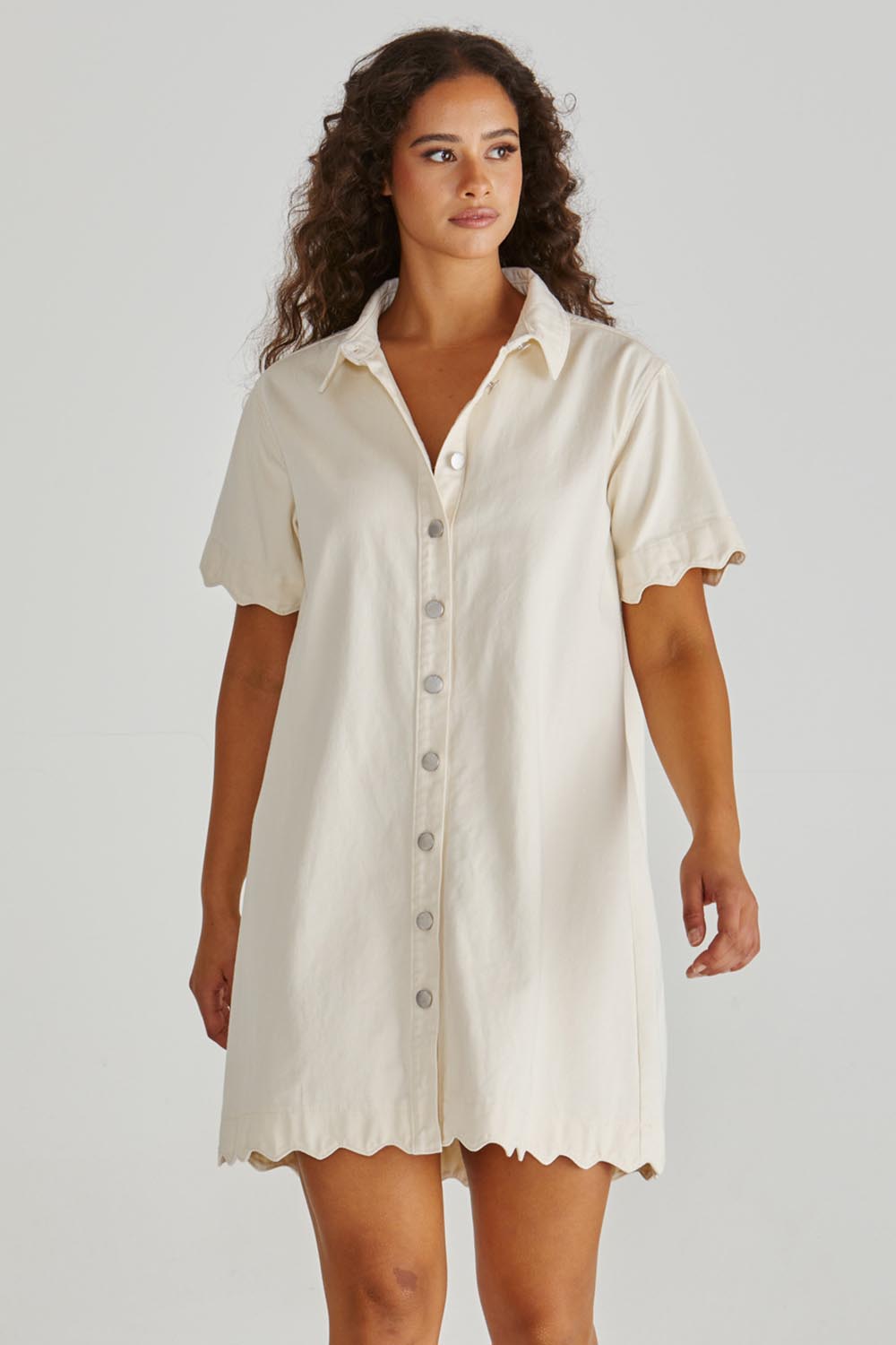 A model wearing the Vale Scalloped Denim Dress in colour Parchment (White), from the front, cropped image