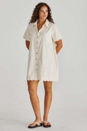 A model wearing the Vale Scalloped Denim Dress in colour Parchment (White), from the front, showing the whole outfit