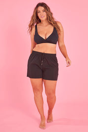 Model Laura wearing size L of the Mid Length Boardshort in colour Black (Black), from the front, showing the whole outfit