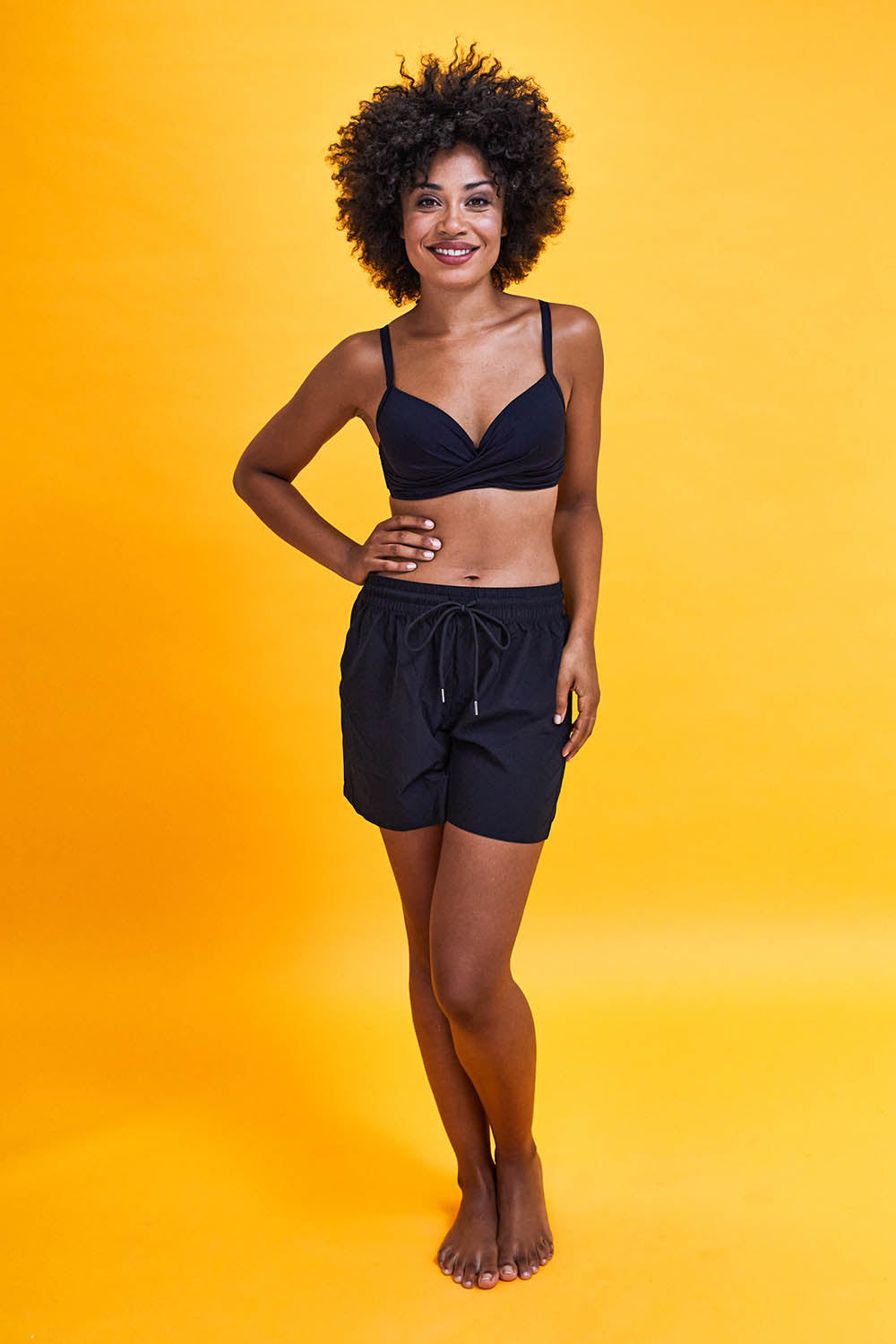 Model Olivia wearing size S of the Mid Length Boardshort in colour Black (Black), from the front, showing the whole outfit
